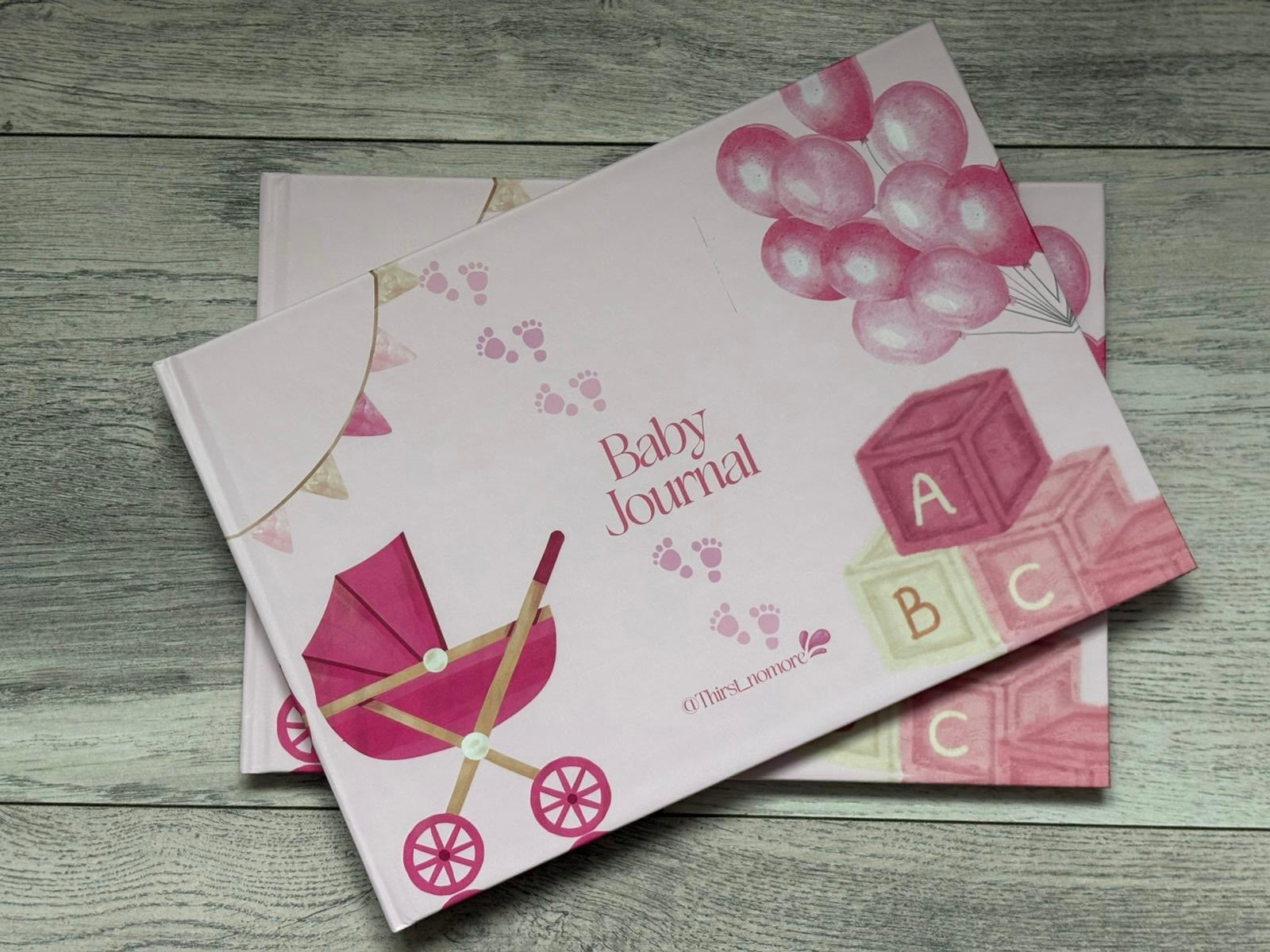 Pink baby journal with illustrations of a stroller, balloons, and letters on a wooden surface